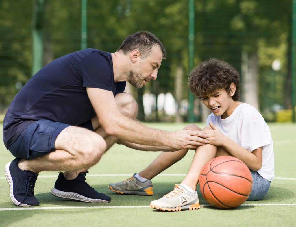 Sports Injury. Handsome PE teacher helping boy with knee trauma after playing basketball